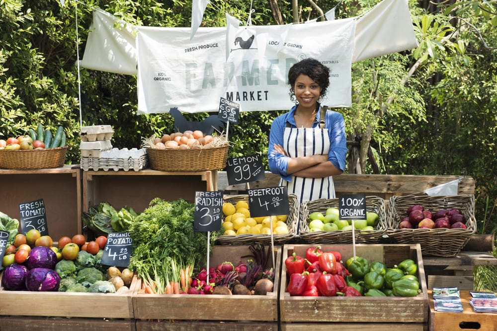 a local farmer standing in front of her market stand at a local farmers' market.