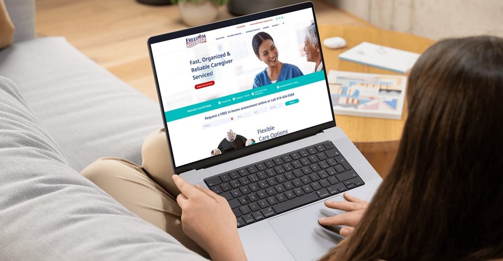 Freedom Caregivers homepage pulled up on laptop held by a woman on a couch
