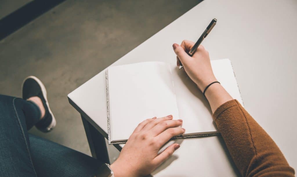 A person's hands writing in a blank notebook on a table, taking notes of questions about Hiring A Columbus Roofer.