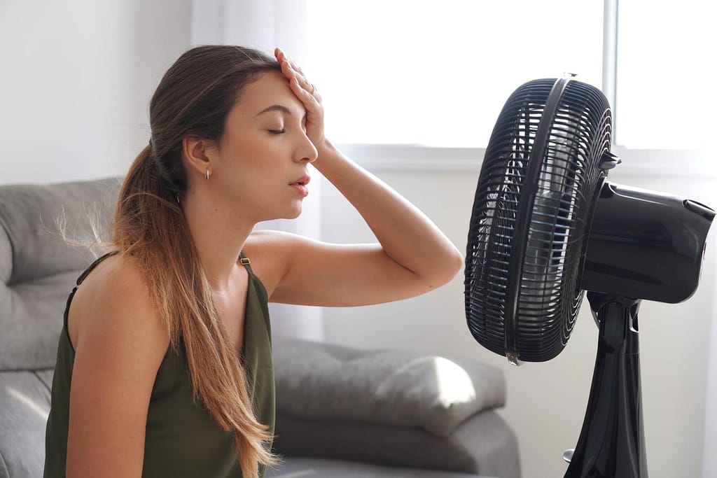Overheated young woman in front of the working fan suffering from summer heat at home. Girl sitting in front of ventilator in hot weather.
