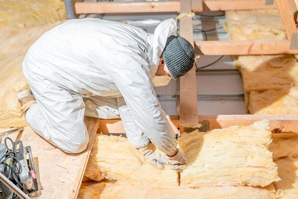 A worker in a white protective suit and gloves is installing attic insulation in a residential attic. Yellow fiberglass insulation batts are being carefully placed between wooden beams, with tools visible on the side. The setting is bright and spacious, highlighting the importance of proper insulation for energy efficiency.