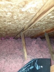 An attic space showing a layer of pink insulation material spread across the floor, with wooden beams and an OSB board ceiling visible, illustrating a potential DIY insulation project.