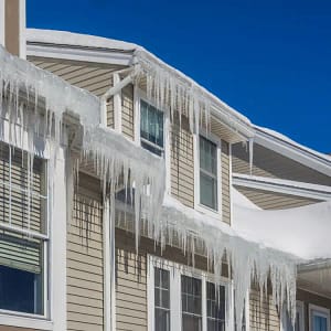 Residential home with snow-covered roof edges and large ice dams hanging from the gutters and eaves against a clear blue sky.