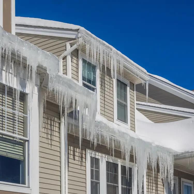 Residential home with snow-covered roof edges and large ice dams hanging from the gutters and eaves against a clear blue sky.